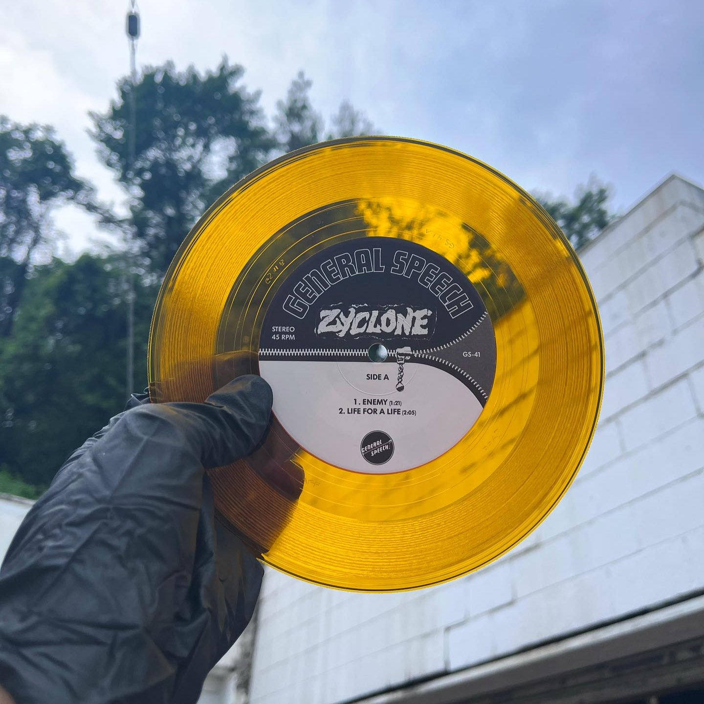 Clear orange vinyl record held by a person outdoors with trees and building in the background