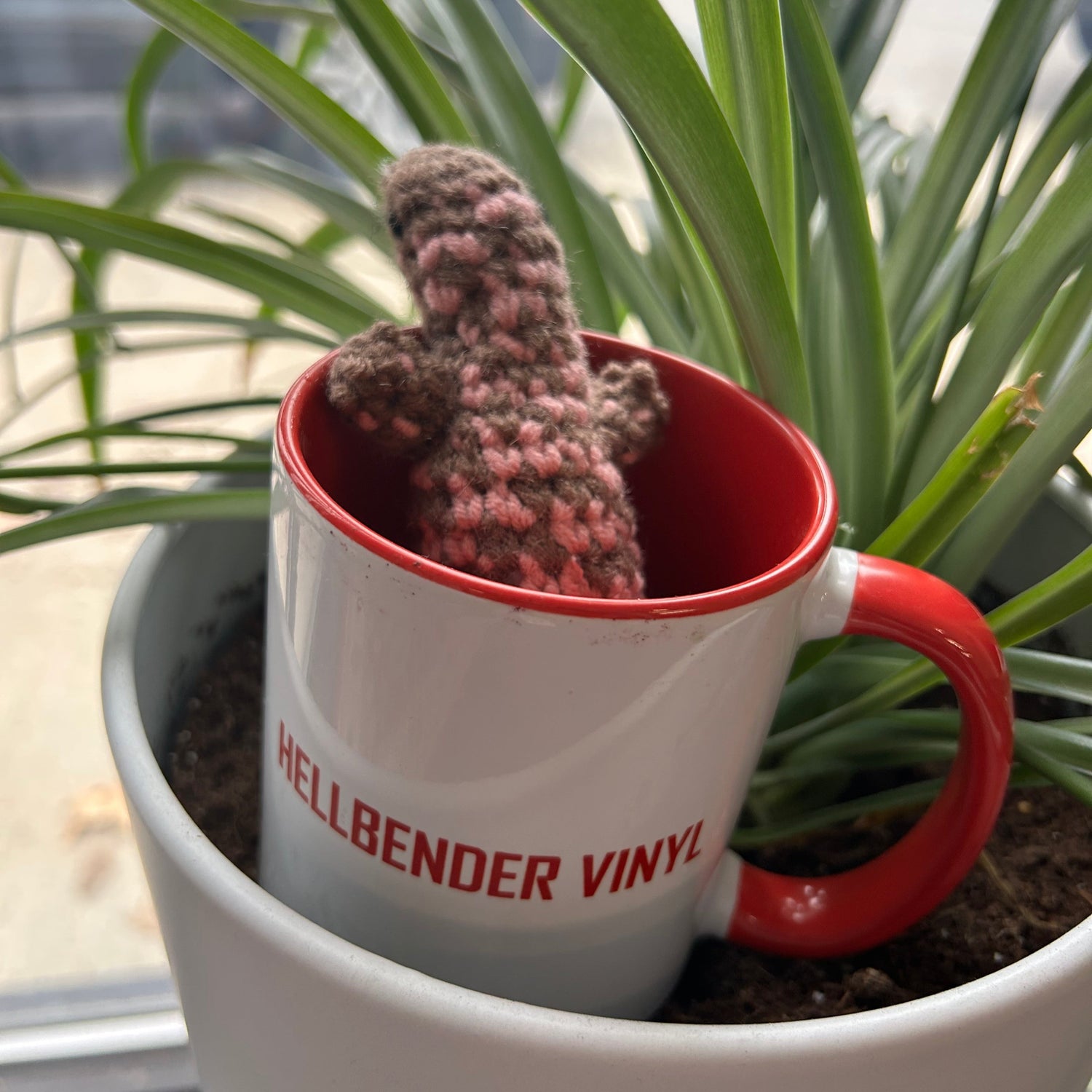 Hellbender Vinyl mug with crocheted Hellbender salamander inside a potted plant on a windowsill